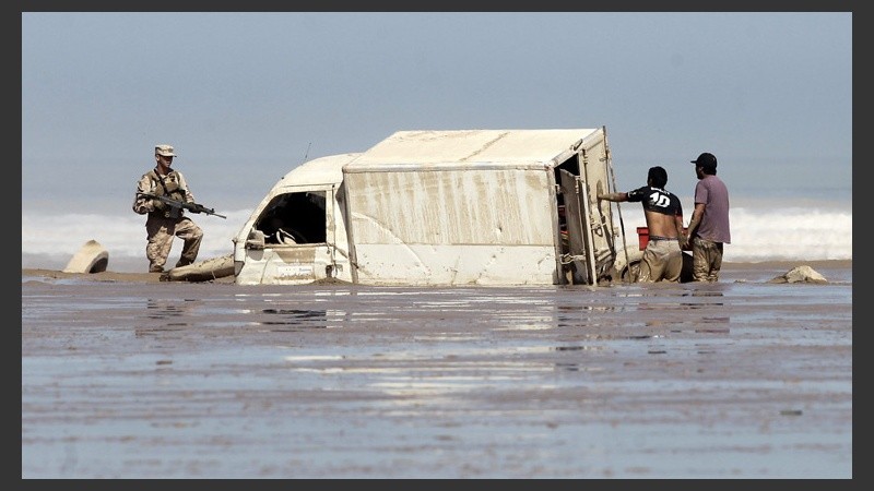 Dos hombres descargan mercancía de un vehículo varado en la playa de Charañal.