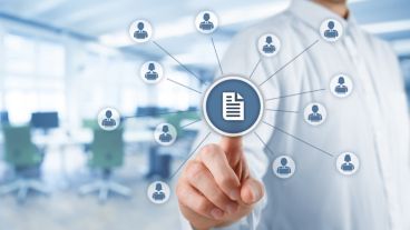 Corporate data management system (DMS) and document management system concept. Businessman click (or publish) on document connected with corporate users working on notebooks with access rights. Wide banner composition with office in background.